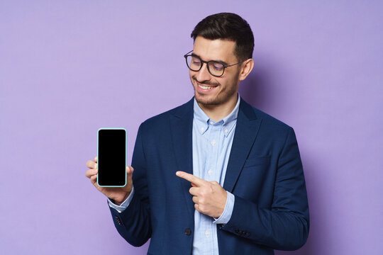 Young business man in jacket, shirt and eyeglasses holding smartphone with blank screen, copy space included, pointing to it, advising high quality finance app, isolated on purple background