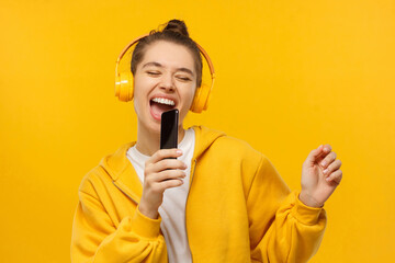 Karaoke online app. Young woman singing out loud in phone as mic, wearing wireless headphones, having fun to favorite song, isolated on yellow background