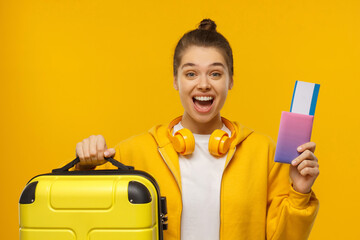Young teenage girl wearing hoodie and wireless headphones around neck, holding travel suitcase and...