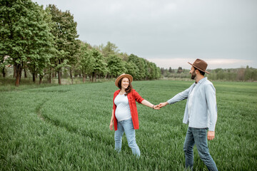 Fototapeta premium Pregnant woman with her man having fun together, walking on the greenfield. Happy couple expecting a baby, young family concept