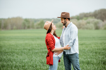 Portrait of a pregnant woman with her boyfriend dressed casually with hats standing together on the greenfield. Happy couple expecting a baby, young family concept