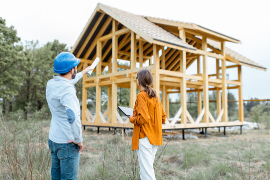 Builder with a female client near the wooden house structure on the construction site outdoors. Building and designing wooden frame house concept