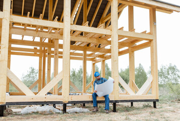 Builder in blue overalls and hard hat with blueprints on the construction site, building wooden frame house