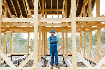 Fototapeta premium Builder in blue overalls and hard hat on the construction site, building wooden frame house
