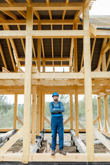 Naklejka na ściany i meble Builder in blue overalls and hard hat on the construction site, building wooden frame house