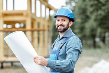 Portrait of a confident builder in blue overalls and hard hat standing with blueprints on the...