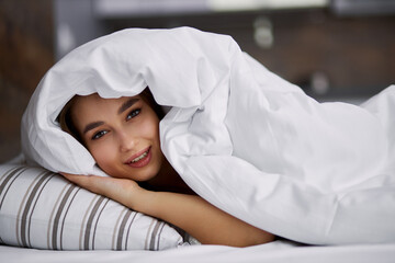 young caucasian woman lie under blanket, she spends her morning on bed in weekends. look at camera