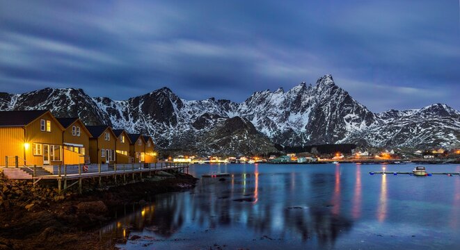 A panoramic evening shot of classic yellow rorbu reflecting the water under cloudy sky and snowy mountain in Ballstad, Lofoten island, Norway