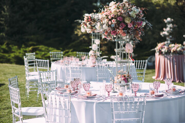 Banquet table on a green lawn. Rack and cutlery. Floral arrangement of pink flowers. Transparent banquet chairs. On the table is a white tablecloth.