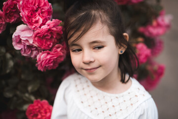 Closeup portrait of cute smiling kid girl 4-5 year old posing in blooming pink roses. Summer season. Childhood.