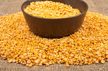 Yellow split peas in a ceramic bowl and grits scattered on a background of coarse-textured burlap. Traditional cereals for making soups and porridge. Close up.