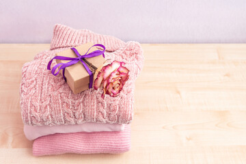 Stack of winter or autumn womens clothes, gift box and flowers. Pile of colorful knitted cozy warm pink sweaters or pullover on wooden shelf.