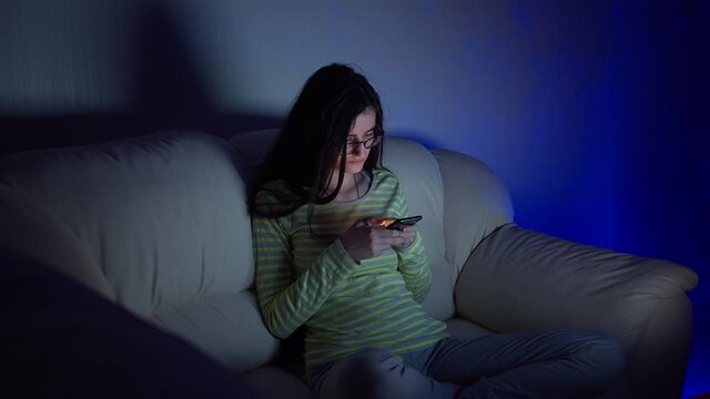 Cute teen girl in glasses sitting on the couch with his legs crossed and reading a conversation on your phone