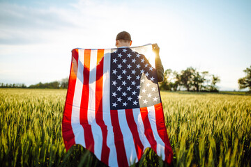 Young man wearing green shirt and cap stands wrapped in the american flag at the green wheat field. Patriotic boy celebrates usa independence day on the 4th of July with a national flag in his hands.