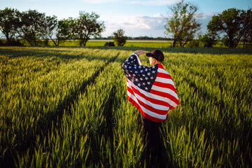 Young man wearing green shirt and cap stands wrapped in the american flag at the green wheat field....