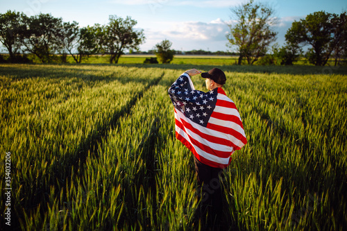 Young man wearing green shirt and cap stands wrapped in the american flag at the green wheat field. Patriotic boy celebrates usa independence day on the 4th of July with a national flag in his hands.