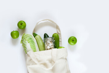 Reusable canvas bag with green apples, pumpkin seeds in a glass jar, fresh vegetables on a white background. Concept zero waste shopping, healthy food, vegetable food. Flat lay, top view, copy space.