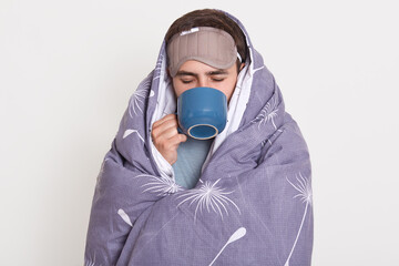 Indoor shot of unshaven male spending time at home in morning, man wrapped in blanket holding blue...