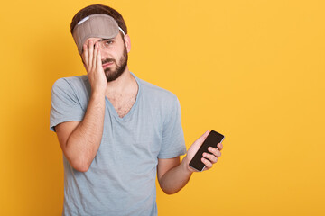 Unshaven dark haired male dresses casual gray t shirt and sleeping mask, holding smart phone in hand, covering half of his face with palm, standing against yellow wall. Copy space for advertisement.