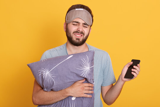 Tired sleepy man with closed eyes wearing blindfold and t shirt, embracing gray pillow, unshaven guy with smart phone in hand, wants to go to bed, posing isolated over yellow background.