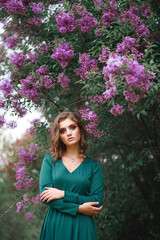 Fashionable Portrait of a young beautiful beautiful girl posing against a background of lilac bushes in bloom in a green dress