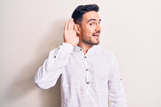 Young handsome man with beard wearing casual t-shirt standing over white background smiling with hand over ear listening and hearing to rumor or gossip. Deafness concept.