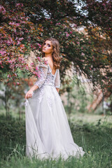 Obraz na płótnie Canvas A beautiful romantic girl in a white delicate dress stands near a Bush of pink flowers and touches a branch with flowers