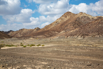 Dry barren landscape with bushes in front of multi-colored hills under cloudy sky in Oman's Sharqiyah governorate