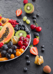 Fresh raw organic summer berries and exotic fruits in white plate on black background. Pineapple, papaya, grapes, nectarine, orange, apricot, kiwi, pear, lychees, cherry and physalis. Close up