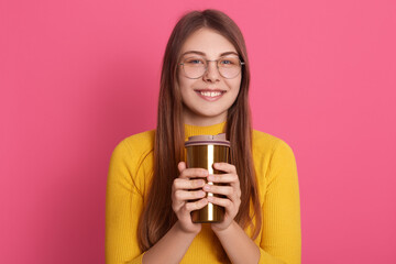 Portrait of relaxed young woman standing indoor and holding cup of coffee, looking directly at camera with charming smile, attractive female wearing yellow shirt and glasses, enjoys hot beverage.