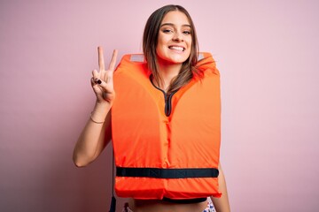 Young beautiful brunette woman wearing orange lifejacket over isolated pink background smiling with happy face winking at the camera doing victory sign. Number two.