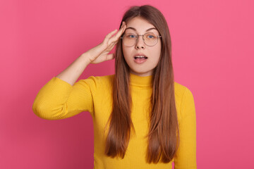 Closeup portrait of female remembers something important, standing with opened mouth and keeping hand near her temple, winsome lady dresses casual attire and glasses, poses against rosy wall.