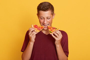 Image of delighted hungry young man eating two slices of pizza, being fond of junk food, wearing...