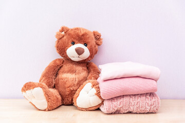 Teddy bear sits near stack of winter or autumn womens clothes. Pile of rose knitted cozy warm pink sweaters or pullover on wooden table.