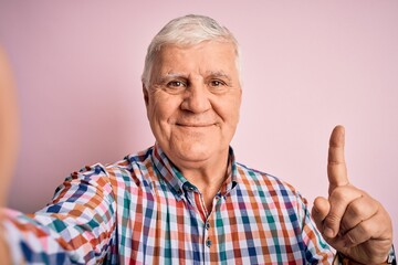 Senior handsome hoary man wearing casual shirt making selfie by the camera surprised with an idea or question pointing finger with happy face, number one