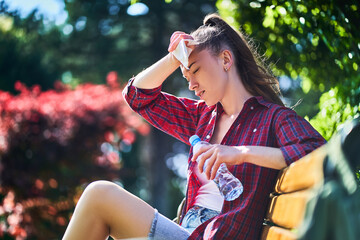 Tired sweating woman wipes her forehead with a napkin and holds cold water bottle in a park in...