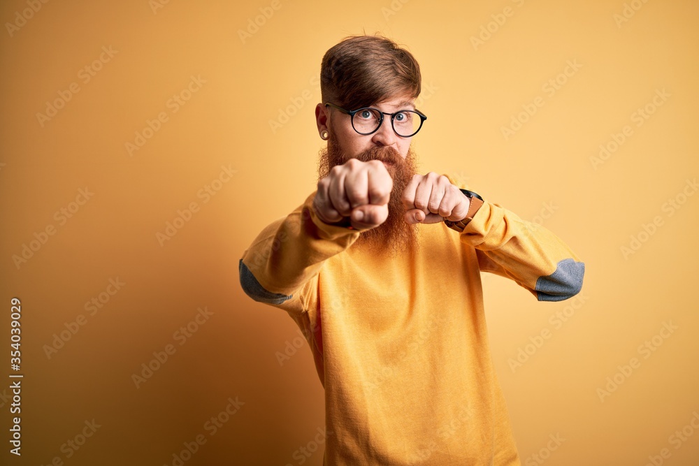 Poster handsome irish redhead man with beard wearing glasses over yellow isolated background punching fist 