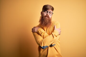 Handsome Irish redhead man with beard wearing glasses over yellow isolated background Hugging oneself happy and positive, smiling confident. Self love and self care