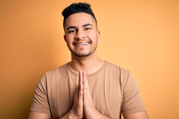 Young handsome man wearing casual t-shirt standing over isolated yellow background praying with hands together asking for forgiveness smiling confident.