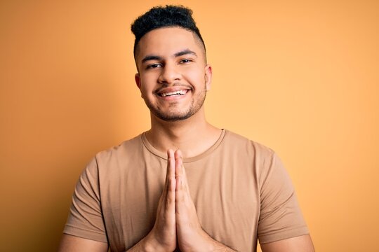Young handsome man wearing casual t-shirt standing over isolated yellow background praying with hands together asking for forgiveness smiling confident.