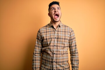 Young handsome man wearing casual shirt standing over isolated yellow background angry and mad screaming frustrated and furious, shouting with anger. Rage and aggressive concept.