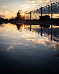 Reflection of the setting sun and clouded skies in the puddle along the fence