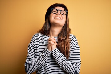 Young beautiful brunette woman wearing french beret and glasses over yellow background begging and...