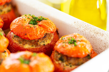 Close-up on French stuffed tomatoes with meat, bread crumbs, and herbs in a white ceramic dish, aside a bottle of olive oil.