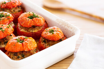 Mediterranean stuffed tomatoes with meat, bread crumbs, and herbs in a white oven dish, aside a white napkin and a wooden serving spoon on an oak wood table.
