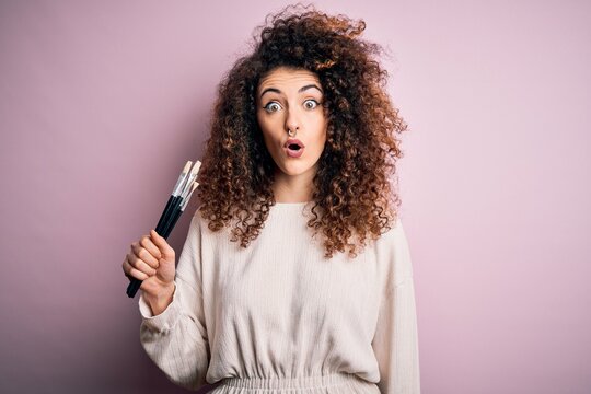 Young beautiful woman with curly hair and piercing applying cosmetic using paint brushes scared in shock with a surprise face, afraid and excited with fear expression