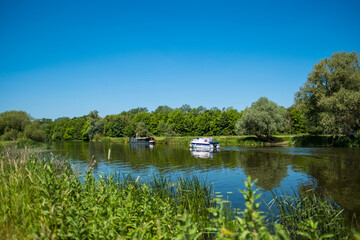 Fototapeta na wymiar Idyllic scenic view of both a narrow boat and motor launch seen in an inland waterway in the UK. The wide expanse of the river is seen as well as the dense foliage and distant woodlands.