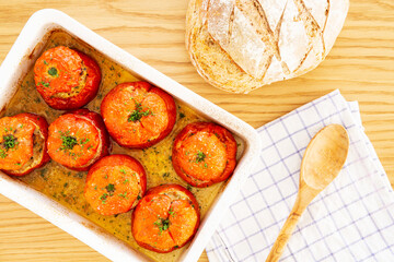 Mediterranean stuffed tomatoes with meat, bread crumbs, and herbs in a white oven dish, aside a kitchen towel,, a bread loaf and a wooden serving spoon on an oak wood table. Flat Lay, top view.