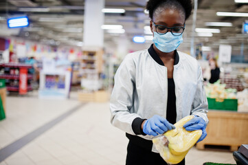 Fototapeta na wymiar African woman wearing disposable medical mask and gloves shopping in supermarket during coronavirus pandemia outbreak. Black female choose bananas at epidemic time.