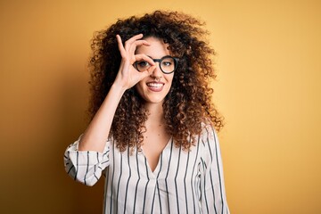 Young beautiful woman with curly hair and piercing wearing striped shirt and glasses doing ok gesture with hand smiling, eye looking through fingers with happy face.